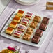 A square white platter with an assortment of pre-cut cheesecake and dessert bars in various flavors.
