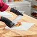 A person wearing black gloves is wrapping a sandwich with a sheet of Choice 16" x 16" insulated deli wrap paper on a wooden countertop.