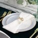 A round pearl white plastic plate with a gold band, set on a table with a white napkin and gold utensils.