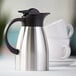 A stainless steel insulated coffee carafe with a black lid and handle.