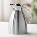 A polished stainless steel insulated carafe with a flip-top lid and handle.