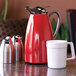 A red stainless steel insulated coffee carafe with a push-button lid, shown alongside a white mug and two metal dispensers.