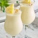 A pair of creamy yellow beverages garnished with pineapple wedges and coconut pieces, served in curved glasses.