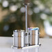 A brushed stainless steel sugar stick holder and creamer set with a central handle.