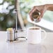 A stainless steel sugar stick holder and creamer set next to a white coffee mug.