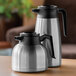 A pair of brushed stainless steel insulated coffee carafes with black handles and lids, one tall and one short.