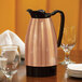A copper stainless steel insulated coffee carafe with a black lid and handle, placed on a dining table.