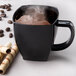 A black square-shaped disposable plastic coffee mug filled with a hot beverage, shown with coffee beans and wafer rolls nearby.