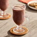 A glass mug filled with a chocolate-colored beverage, likely made from carob powder, placed on a woven coaster.
