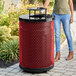 A large, round, red expanded steel outdoor trash can with a rain bonnet lid.