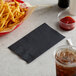 A black 2-ply paper dinner napkin folded in 1/8 style, placed on a table next to food and drinks.