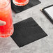 A black 2-ply cocktail napkin placed on a light-colored surface next to a glass of red beverage.