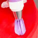 A metal piping tip attached to a piping bag is being used to apply purple icing onto a red surface.