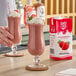 A carton of Island Oasis Strawberry Beverage Mix next to two glasses of strawberry smoothies topped with whipped cream and garnished with strawberries.