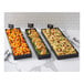A set of three rectangular black metal market trays filled with assorted foods, displayed on a marble countertop.