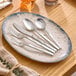A five-piece set of stainless steel flatware, including two forks, a knife, and two spoons, displayed on a ceramic plate.