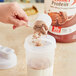 A scoop of chocolate protein powder being poured into a shaker cup with milk, with a container of Premier Protein powder in the background.