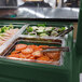 A green Cambro salad bar on a table with trays of tomatoes and vegetables.