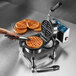 A commercial single cast iron Belgian waffle iron with a waffle being removed using tongs and several cooked waffles on a cooling rack.