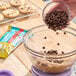 A bowl of cookie dough being mixed with semi-sweet chocolate morsels, with a package of Nestle Toll House Organic Allergen-Free Semi-Sweet Chocolate Morsels and baked cookies in the background.