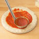 A pizza dough with sauce being spread on it using a spoon.