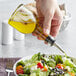A clear glass oil cruet with a stainless steel pourer being used to pour oil onto a salad.