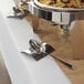 A brushed stainless steel square spoon rest holding a serving spoon on a buffet table.