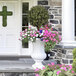 A large artificial potted boxwood ball topiary in a white urn, surrounded by colorful flowers, displayed on a porch.