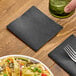A black linen-feel beverage napkin folded into a square, placed on a wooden table.
