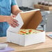 A white, customizable bakery box containing a cake, being closed by a person in a kitchen setting.
