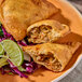 A Philly cheesesteak empanada with a flour shell, served with lime wedges and shredded cabbage on the side.