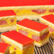 A rectangular layered dessert with a glossy red top, yellow and cream layers, and visible pieces of fruit inside.