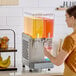 A double-bowl refrigerated beverage dispenser with clear containers holding yellow and red drinks, being used by a person to fill a cup.