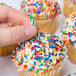 A hand placing a green and white striped birthday candle into a cupcake topped with colorful sprinkles, with other similarly decorated cupcakes in the background.