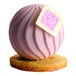 A pink spherical chocolate dessert with a textured surface, decorated with a white and pink square, sitting on a round cookie base.