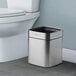 A stainless steel square wastebasket with a 2-gallon capacity placed next to a toilet.
