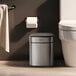 A stainless steel square wastebasket with an automatic lid, placed next to a toilet in a bathroom.