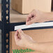 A person applying a white slip strip label holder to the edge of a metal shelving unit.