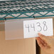 A clear 3" x 5" hanging label holder attached to a wire shelf, with a hand inserting a paper label.