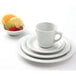 A white Tuxton tall cup on a saucer with a bowl of fruit including strawberries and lime slices.