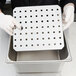 A person in gloves holding a stainless steel tray with holes in it.