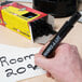 A large black chisel tip permanent marker labeled 'MARKS-A-LOT' being used to write on paper, with a box of similar markers in the background.