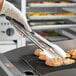 A person in gloves using Tablecraft 2012 12" Ergonomic Locking Tongs to cook food on a grill.