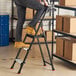 A three-step folding project step stool with a tray being used in a warehouse setting.