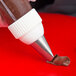 A metal curved petal piping tip attached to a piping bag dispensing chocolate frosting onto a red surface.