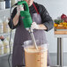 A green heavy-duty immersion blender being used to mix a large container of liquid in a commercial kitchen.