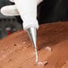 A metal piping tip attached to a piping bag is being used to decorate a chocolate-frosted cake.