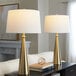 A pair of gold cone-shaped table lamps with white linen drum shades.