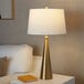 A gold table lamp with a conical base and a white linen drum shade.