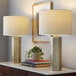 A pair of antique brass table lamps with white linen drum shades placed on a white surface.